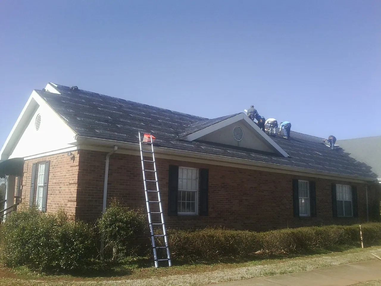 Crew tearing shingles off a brick ranch roof with a ladder set — Metal Roof Repair work by our crew in Wanette