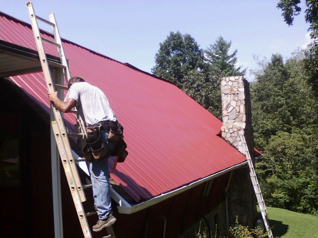 Roofer on a ladder beside a red metal roof and stone chimney — Soffit & Fascia Repair by Roofing Brothers in Wanette